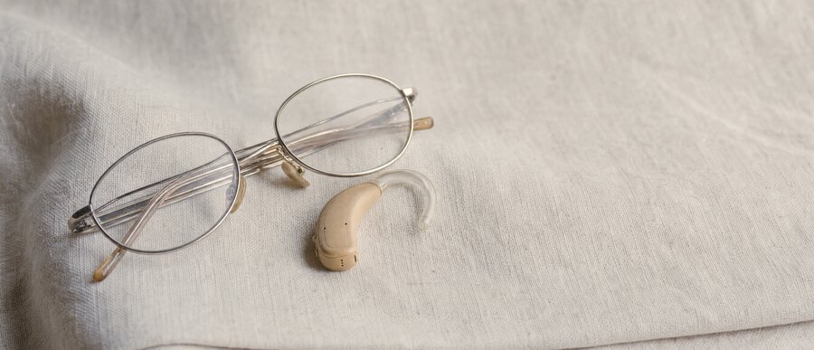 Eine Lesebrille und ein Hörgerät auf hellem Leinen.